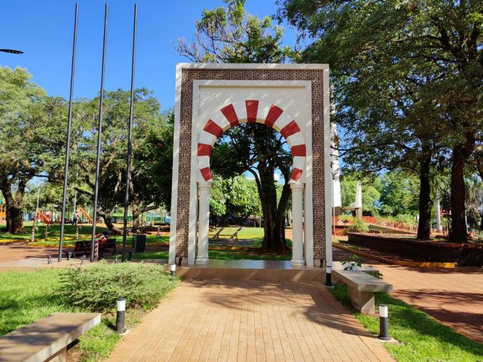 Casi un arbusto todavía, el cedro libanés de destaca sobre el pasto, y al fondo se observa el monumento a la colectividad libanesa, en la Plaza de Armas de Encarnación.